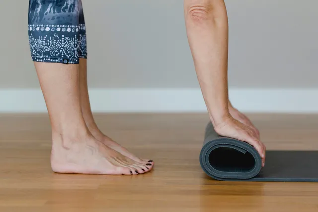 A hand rolls a yoga mat beside bare feet on a wood floor.