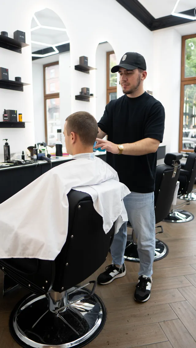 A barber works on a haircut while another customer sits nearby.