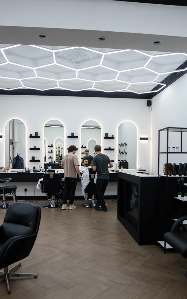 Barber chairs, mirrors and people inside a bright barbershop.