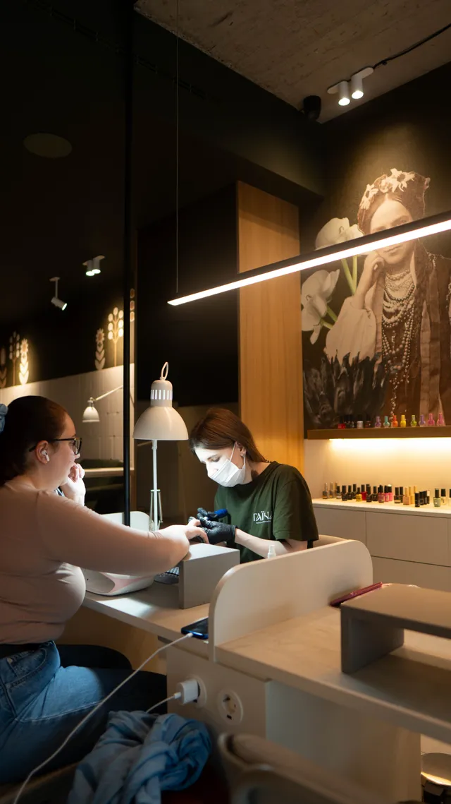 A nail technician and customer sit across a manicure table in warm salon light.