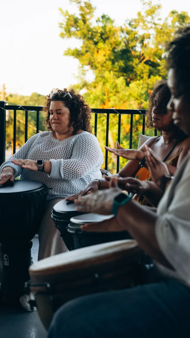 A group plays hand drums together beside sunlit trees.